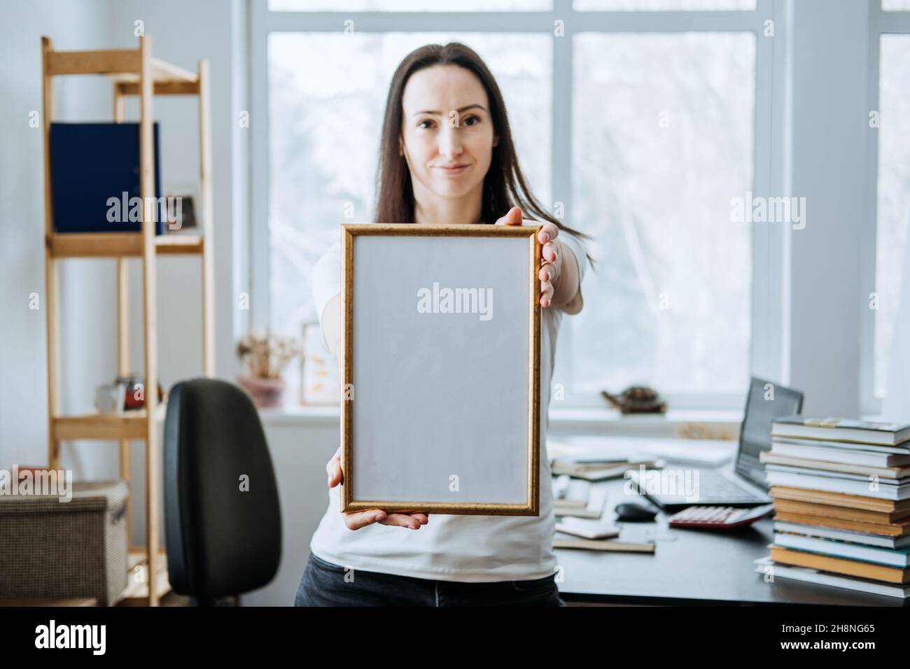 Frame picture, poster, diploma, certificate mockup in female hands. Young brunette woman holding empty blank wooden photo frame on table with laptop Stock Photo