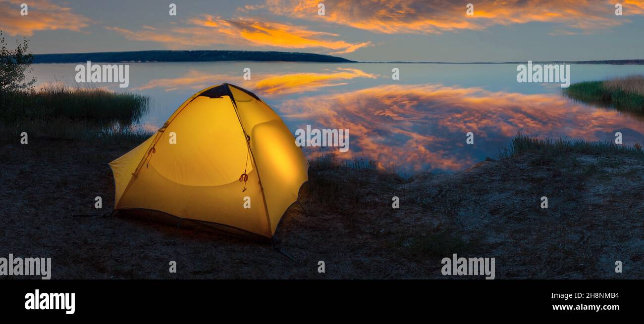Orange tourist tent illuminated from the inside on the lakeside under dramatic sky Stock Photo
