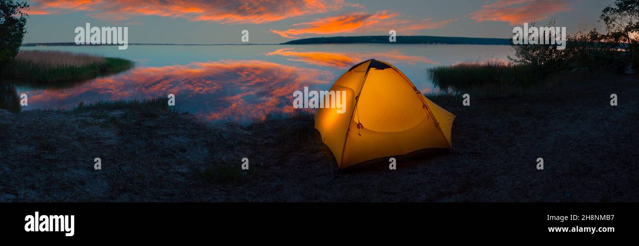 Orange tourist tent illuminated from inside on the lakeside under dramatic sky Stock Photo