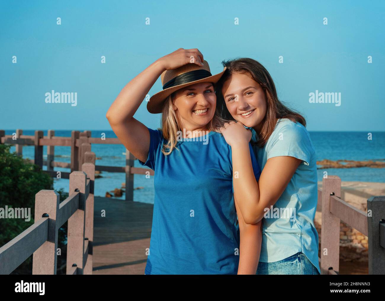 Mother wearing straw hat and teenage daughter standing on pier at sea coast, embracing and smiling, t-shirts mockup. Loving family spending time toget Stock Photo