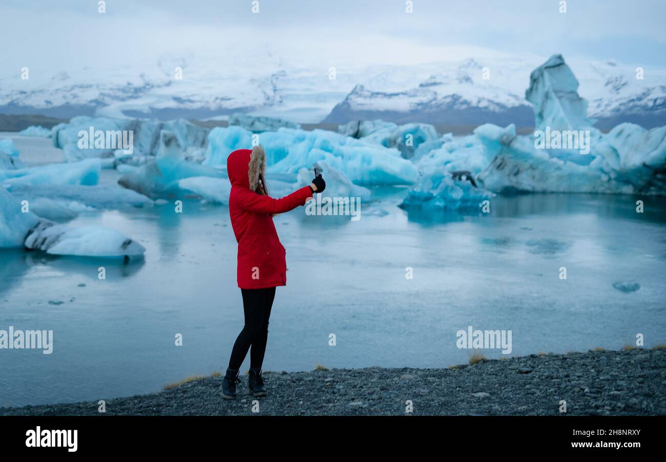 Girl traveler in red jacket with a hood  calling by video internet on smartphone leisure near Jokulsarlon glacial lagoon, Iceland. Stock Photo