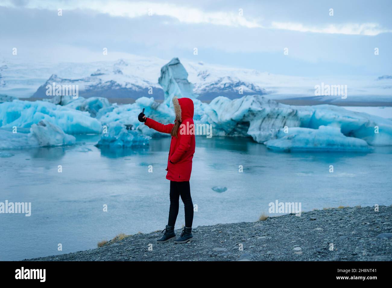Girl traveler in red jacket with a hood  calling by video internet on smartphone leisure near Jokulsarlon glacial lagoon, Iceland. Stock Photo