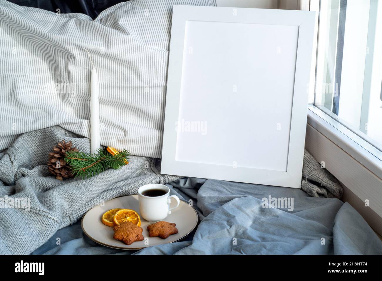 Cozy Christmas breakfast still life scene near the window. Winter interior. Blank white picture frame mockup.Cup of coffee with wholemeal cookies, pil Stock Photo