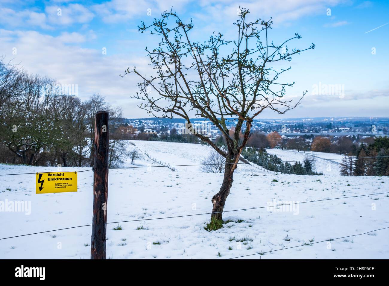 Sign with German text 'Caution Electric Fence' in front of a beautiful snowy winter landscape with blue sky and a few clouds. A small town the back Stock Photo