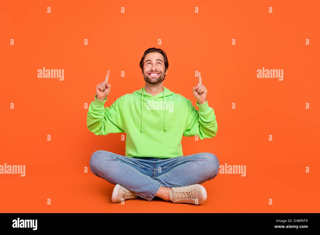 Full body photo of brunet young guy indicate look empty space wear green sweater jeans footwear isolated on orange color background Stock Photo