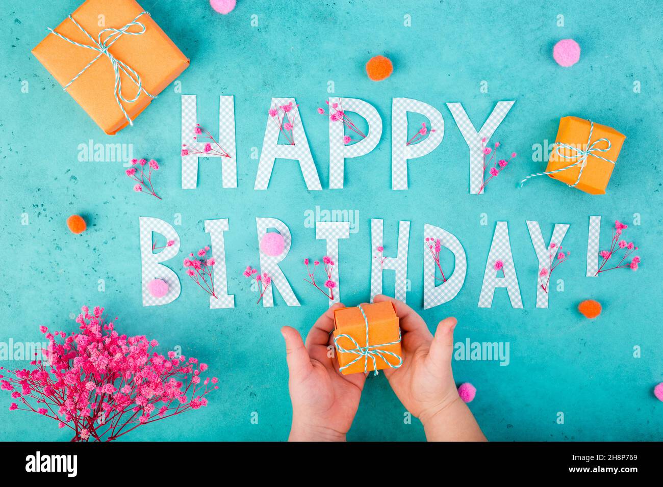 Happy birthday background with letters, orange gifts, pink flowers child hands on turquoise blue background Stock Photo