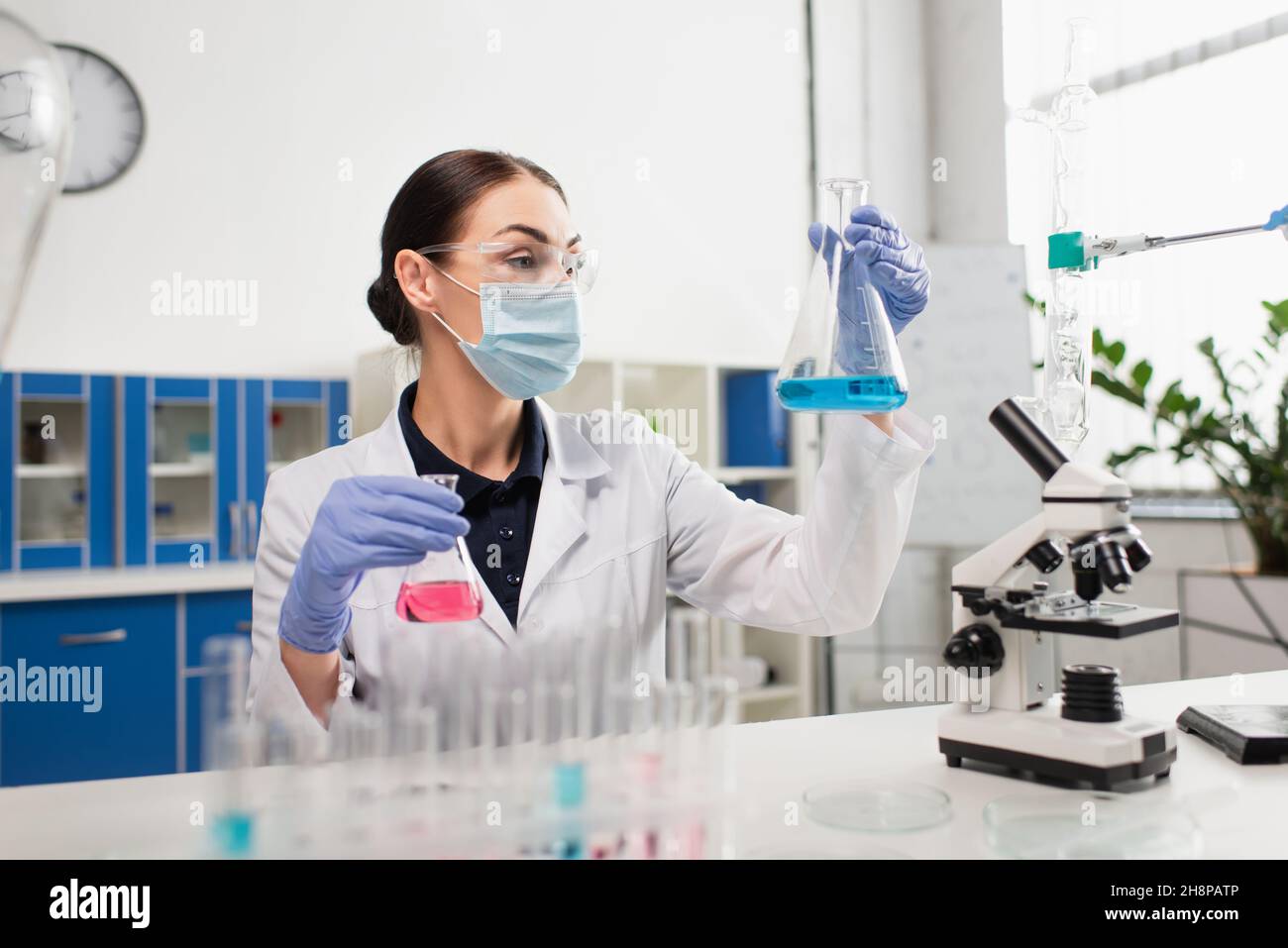 Scientist in safety goggles holding flasks near test tubes and microscope Stock Photo