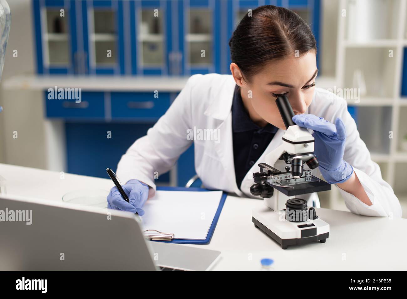 Scientist looking at microscope near clipboard and laptop in lab Stock ...