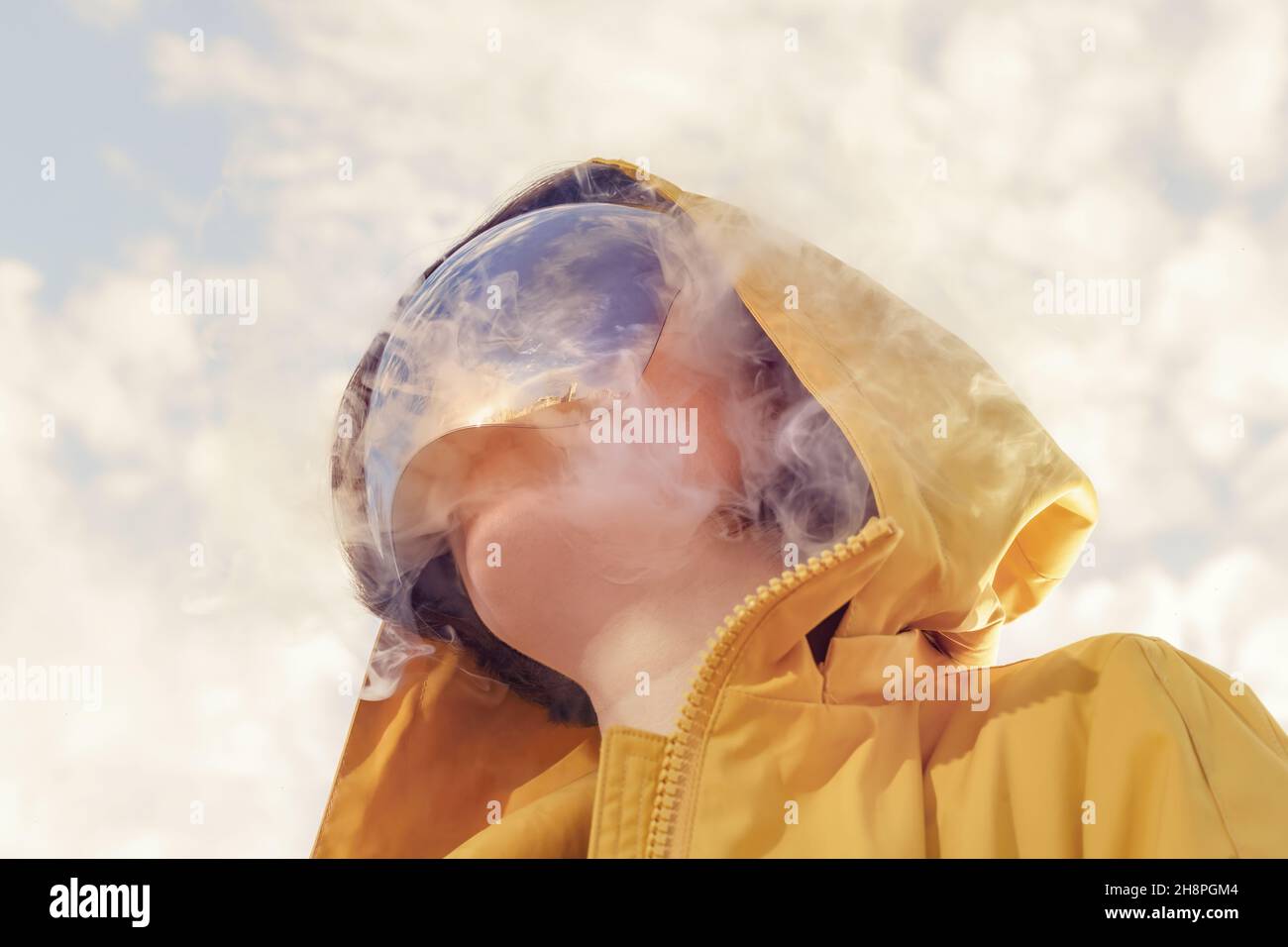 A girl in a yellow raincoat, a mirror mask covering her face and a reflection of the sky in a mask. Futuristic looking surreal portrait Stock Photo