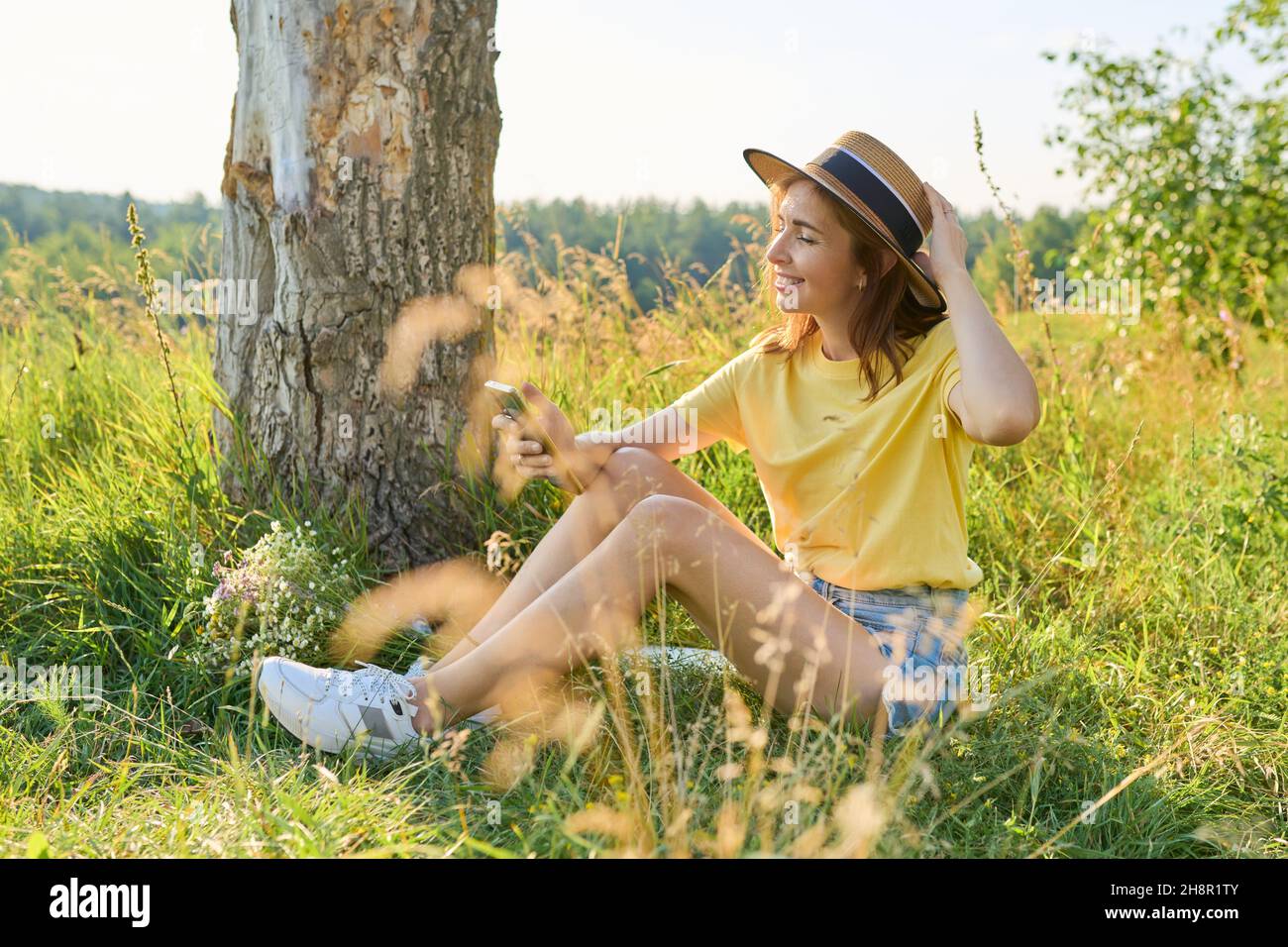 Happy adult woman sitting on meadow, having a rest, using smartphone Stock Photo
