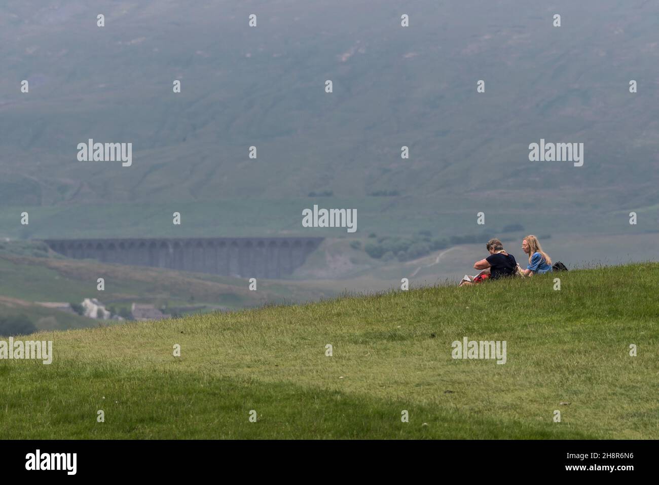 Two hillwalkers resting on the three peaks path sharply focused against a soft focus, heat hazy backdrop of Whernside hill and Ribblehead viaduct. Stock Photo