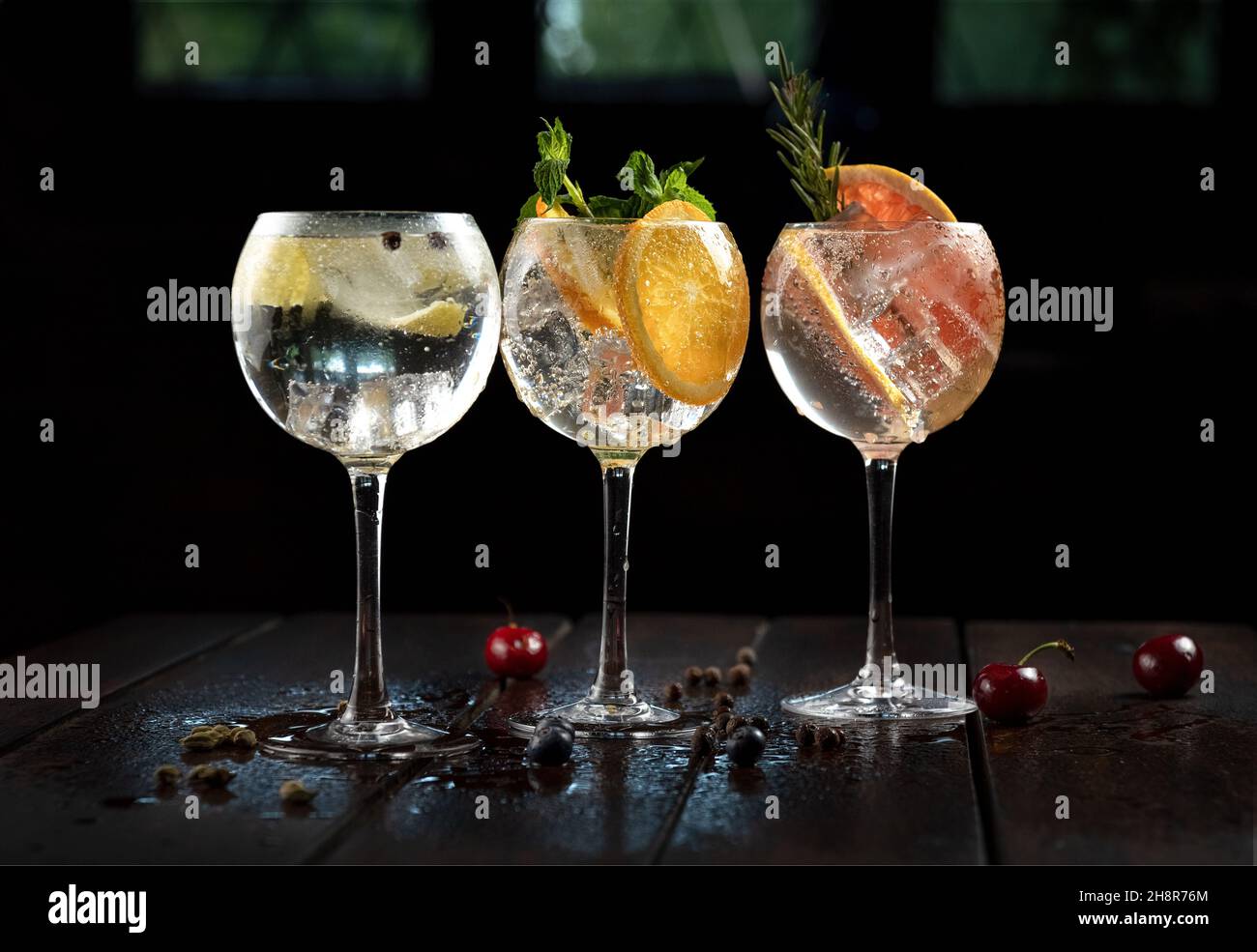 Variedades de bebida alcohólica (tragos de gin and tonic) con limón, pomelo, naranja  y hielo en rústica mesa de madera mojada, adornada con cerezas. Stock Photo