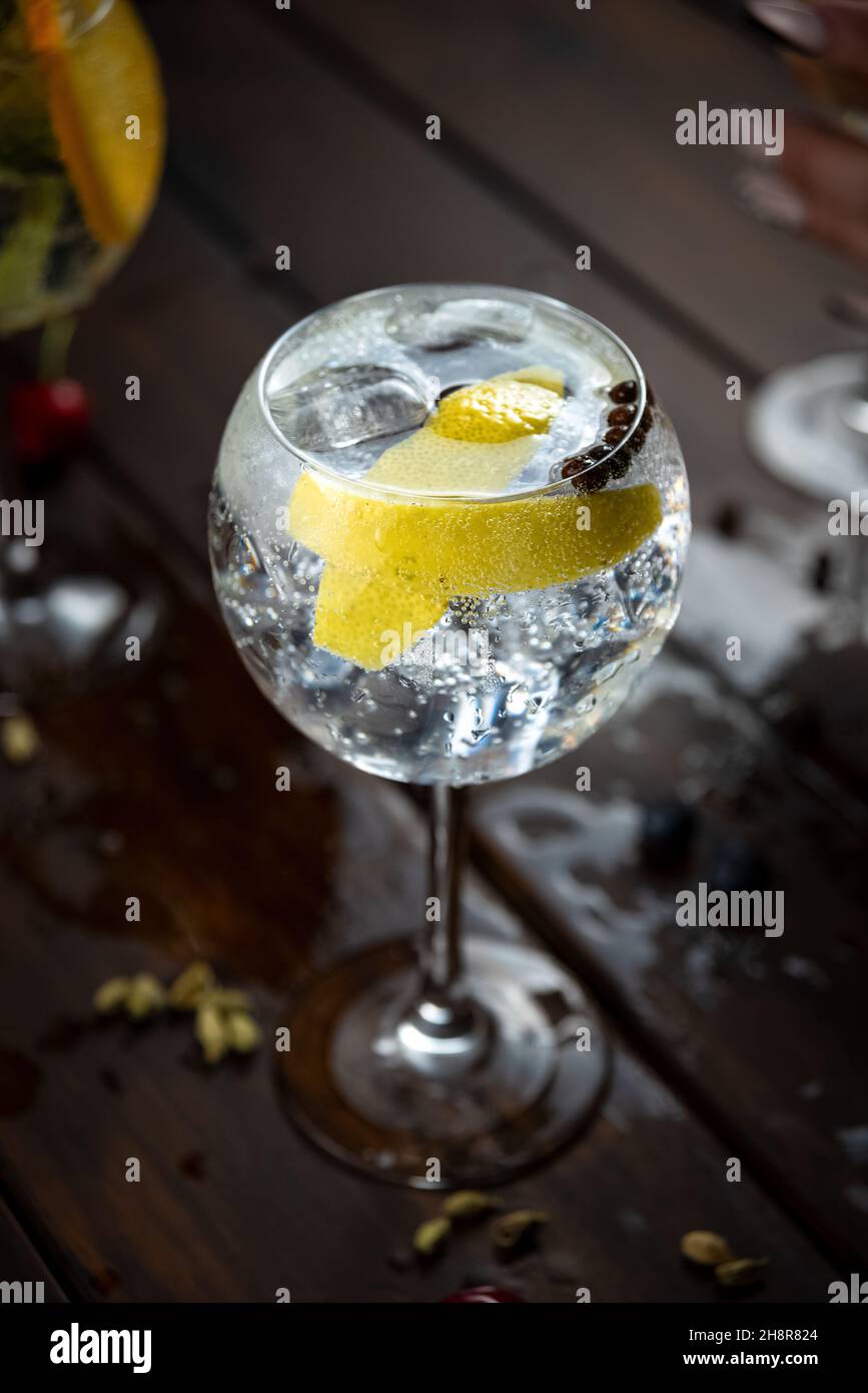 Bebida alcohólica (trago de gin and tonic) con limón y hielo en rústica mesa de madera mojada. Stock Photo