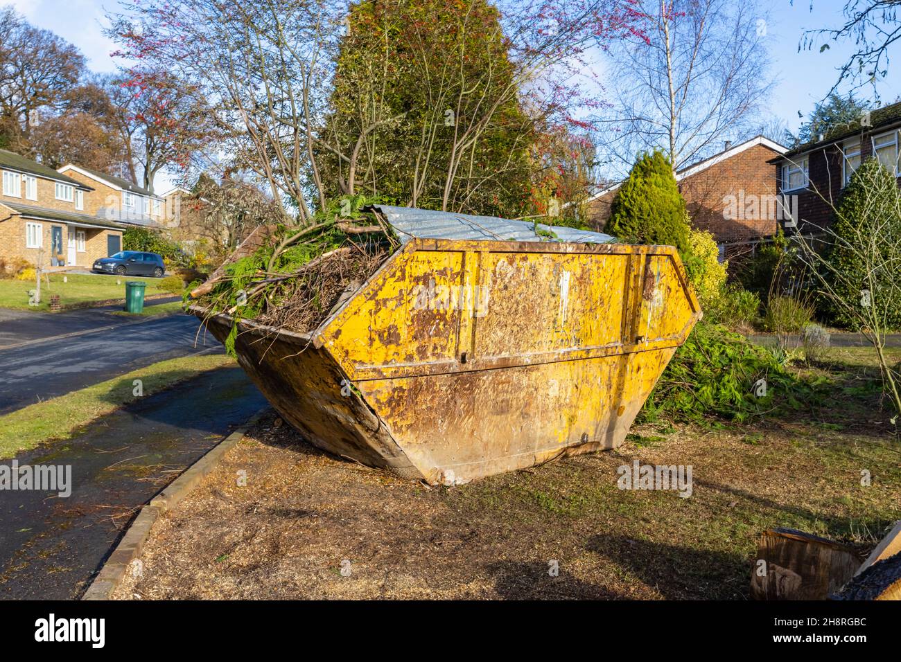 A large rusting, battered waste disposal skip full of branches and garden waste materials on the front garden of a road in Surrey, south-east England Stock Photo