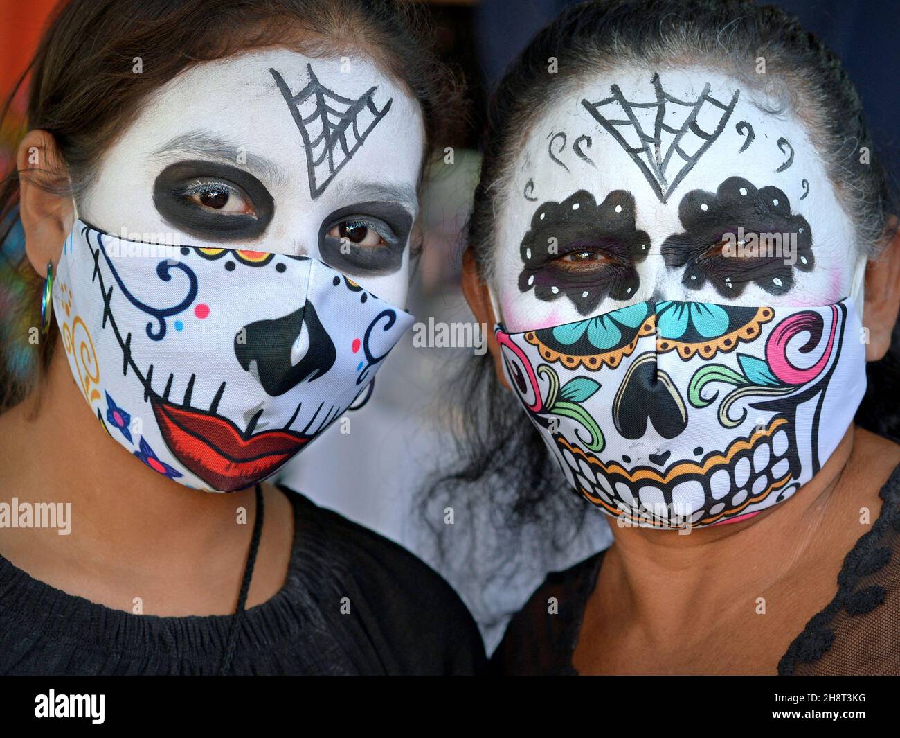Two double-masked Mexican woman with scary Catrina face make-up on the Day of the Dead wear printed cloth face masks during the corona pandemic. Stock Photo