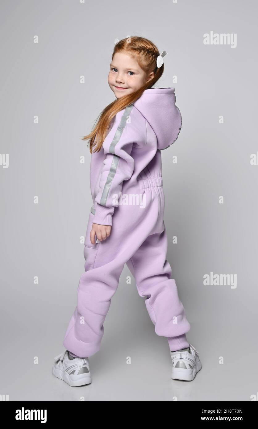 Red-haired kid girl in pink jumpsuit with hood stands back to camera with legs wide apart and turned to camera Stock Photo