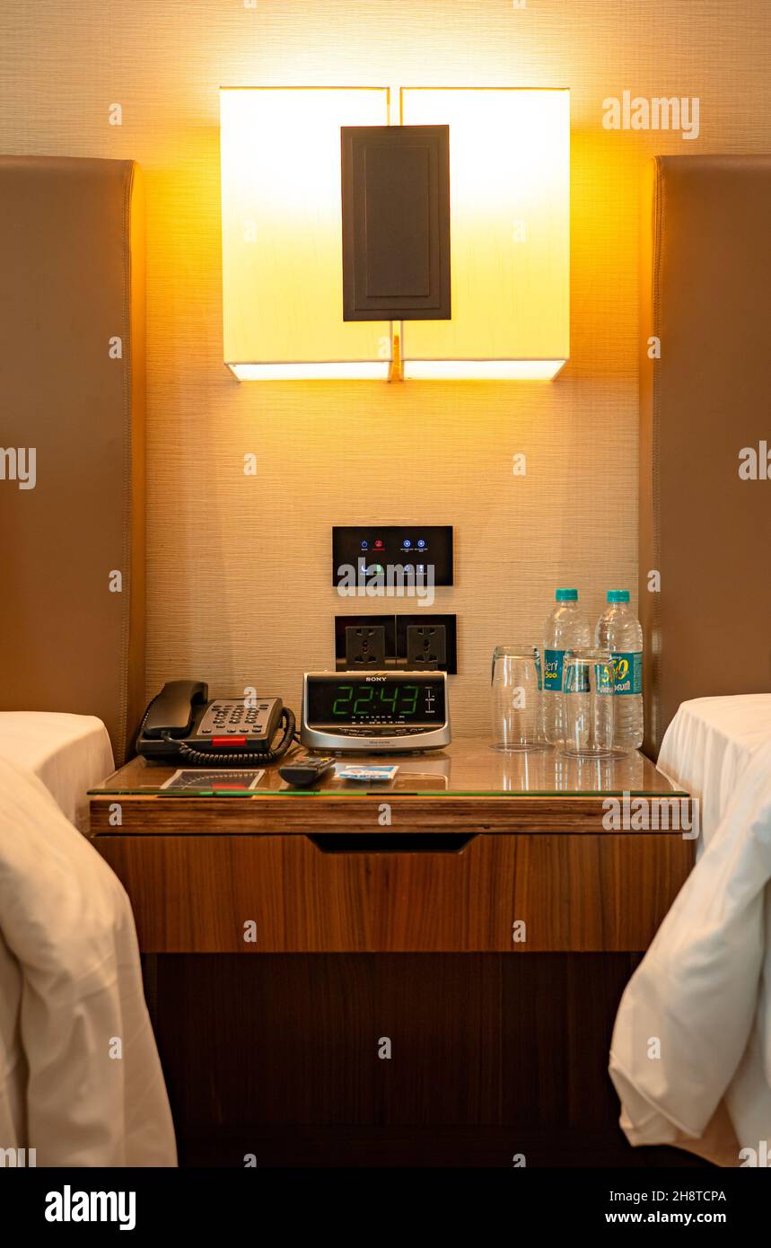 A bedside nightstand table with a telephone, a digital clock, water bottles, and glasses in a luxury hotel room. Stock Photo