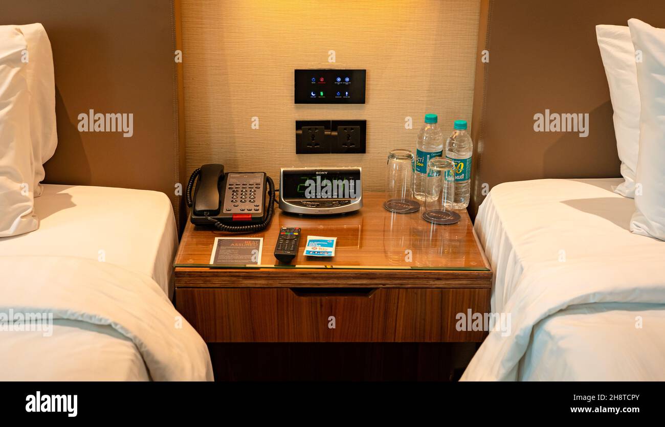 A telephone, digital clock, television remote control, water bottles, and glasses on a small wooden table by the bedside in a modern hotel room. Stock Photo
