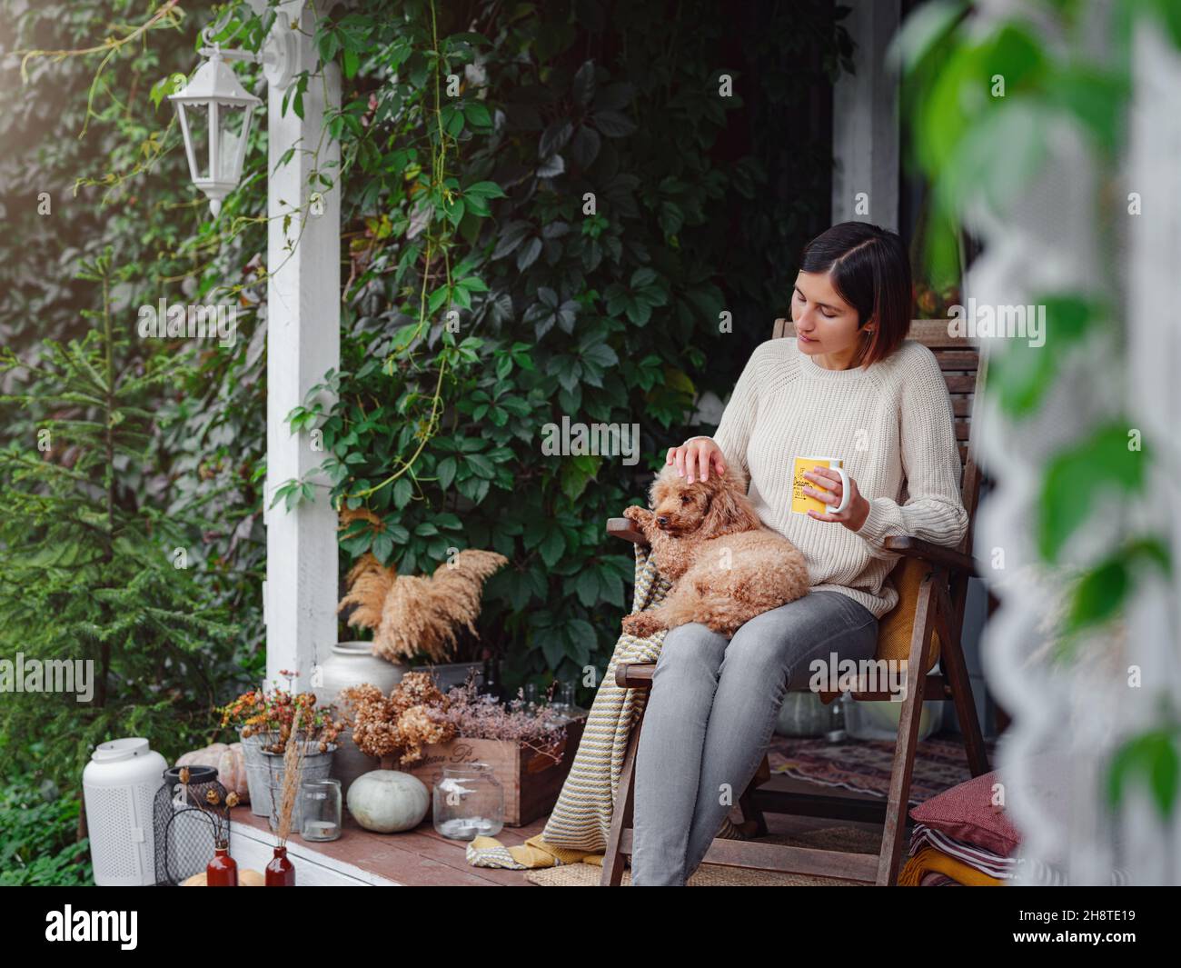 Young asian woman resting outdoors with cup of coffee on porch of country house, chilling outside with poodle dog . autumn lifestyle, leisure free time concept. Copy space Stock Photo