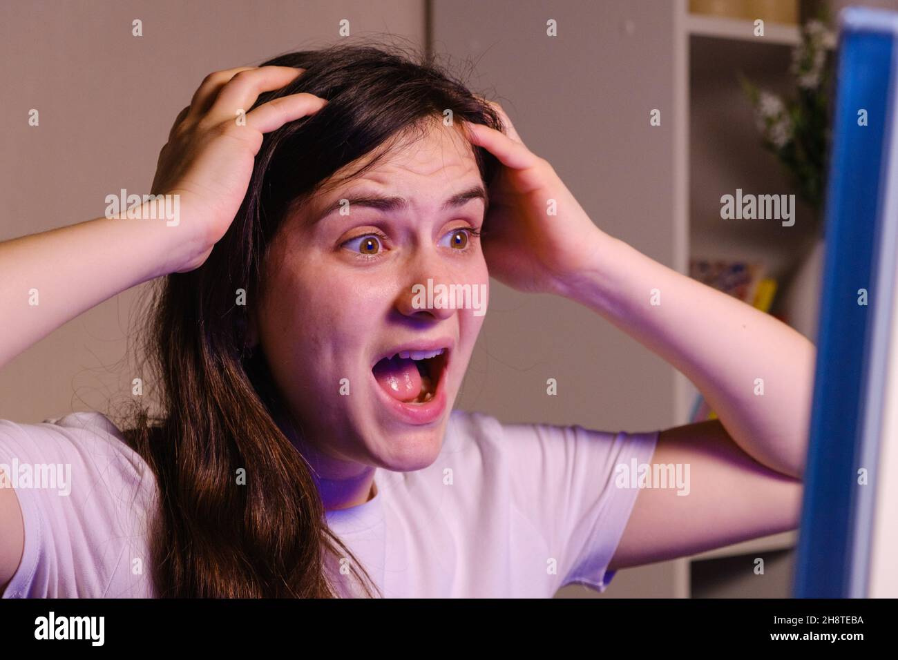 A woman screams and grabs her head with her hands while looking at a TV