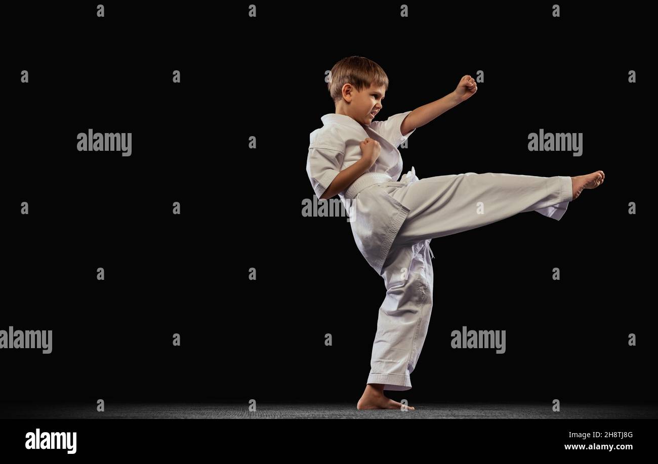 Full-length portrait of little boy, sportsman practising martial art isolated over black background. Side kick Stock Photo