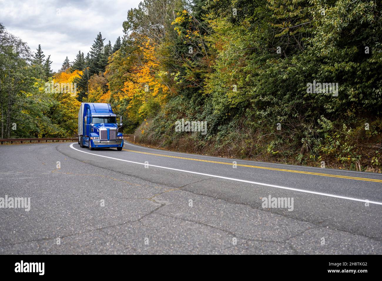 Industrial big rig blue semi truck tractor transporting commercial cargo in refrigerator semi trailer driving on the divided winding highway road with Stock Photo