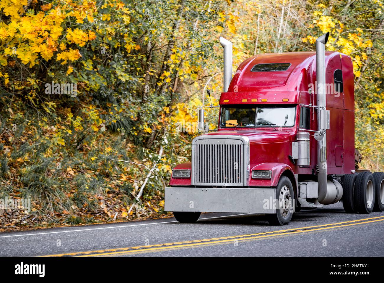 Industrial powerful burgundy big rig semi truck tractor without semi trailer driving on the winding narrow road through the autumn forest in Columbia Stock Photo