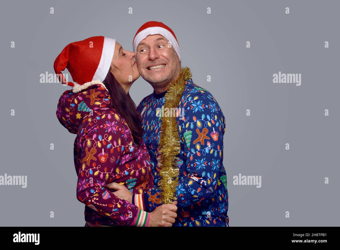Affectionate middle-aged couple in festive holiday themed Christmas outfits embracing and kissing as they celebrate Xmas together over a grey backgrou Stock Photo