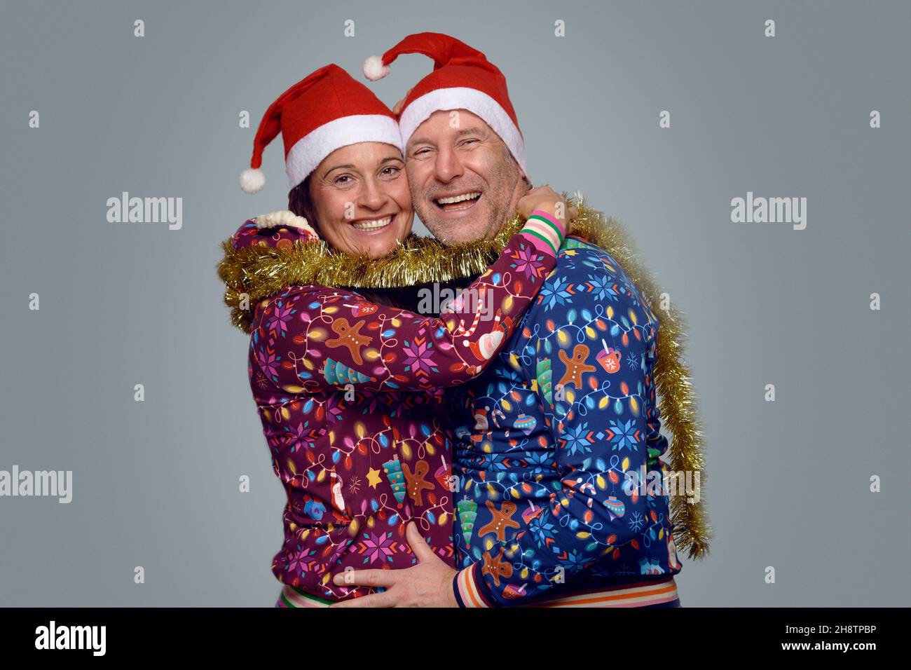 Joyful married couple celebrating Christmas together in festive red Santa hats and themed Xmas tops standing hugging and laughing as they turn to smil Stock Photo