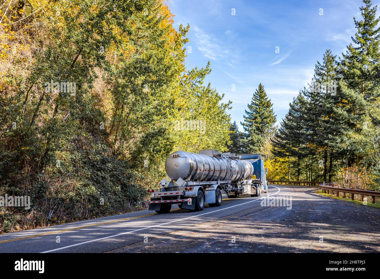 Industrial big rig blue semi truck tractor transporting commercial cargo in armored tank semi trailer driving on the narrow winding highway road with Stock Photo