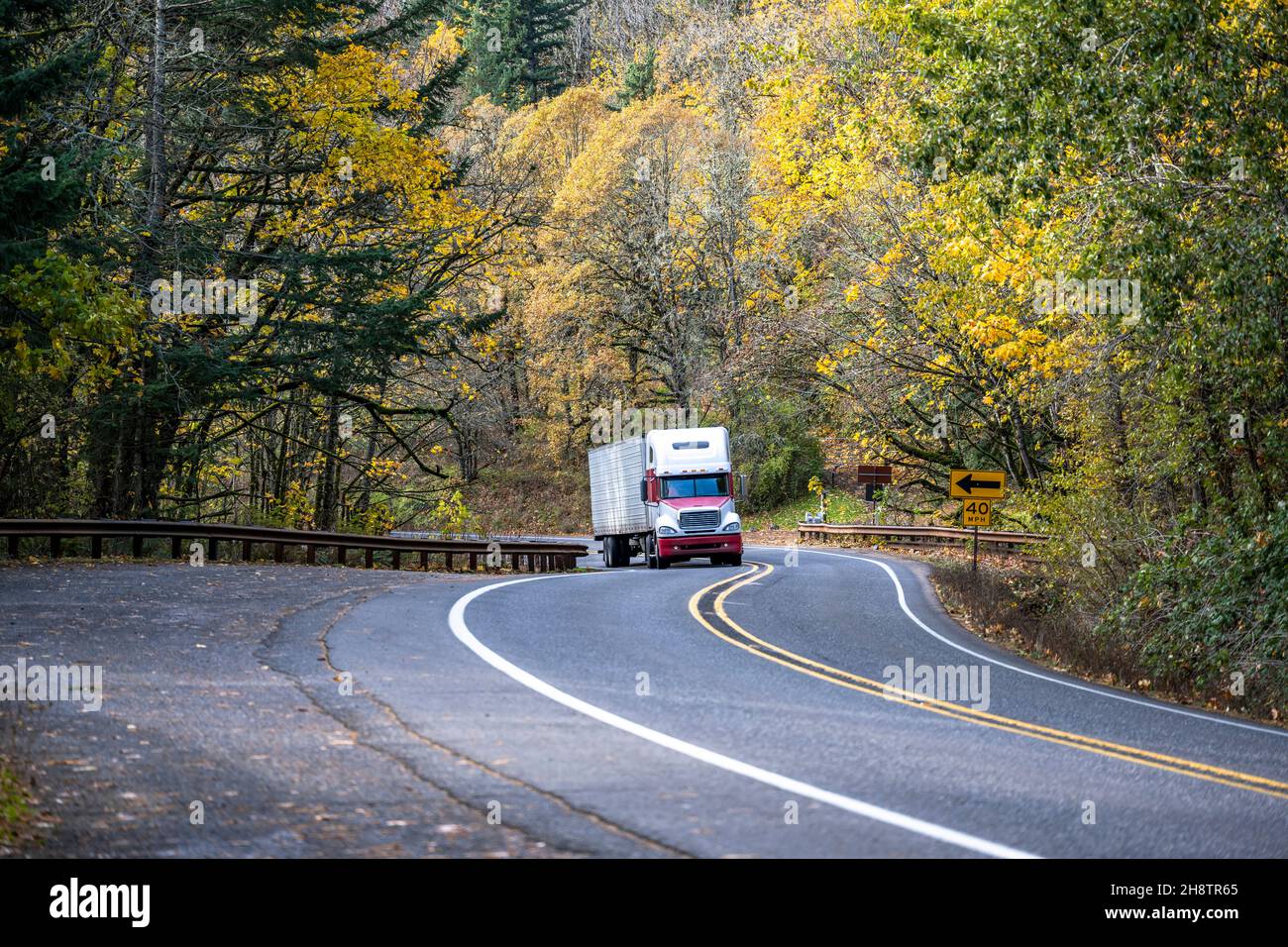 Industrial big rig white and red semi truck tractor transporting commercial cargo in refrigerator semi trailer driving on the narrow winding road with Stock Photo