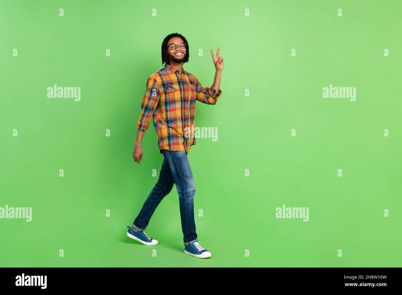 Photo of pretty sweet dark skin guy dressed checkered shirt glasses walking showing v-sign empty space isolated green color background Stock Photo