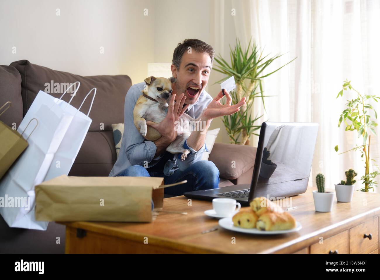 Excited man doing online shopping from a laptop sitting on the sofa with his dog in hand at home. Horizontal composition. Stock Photo