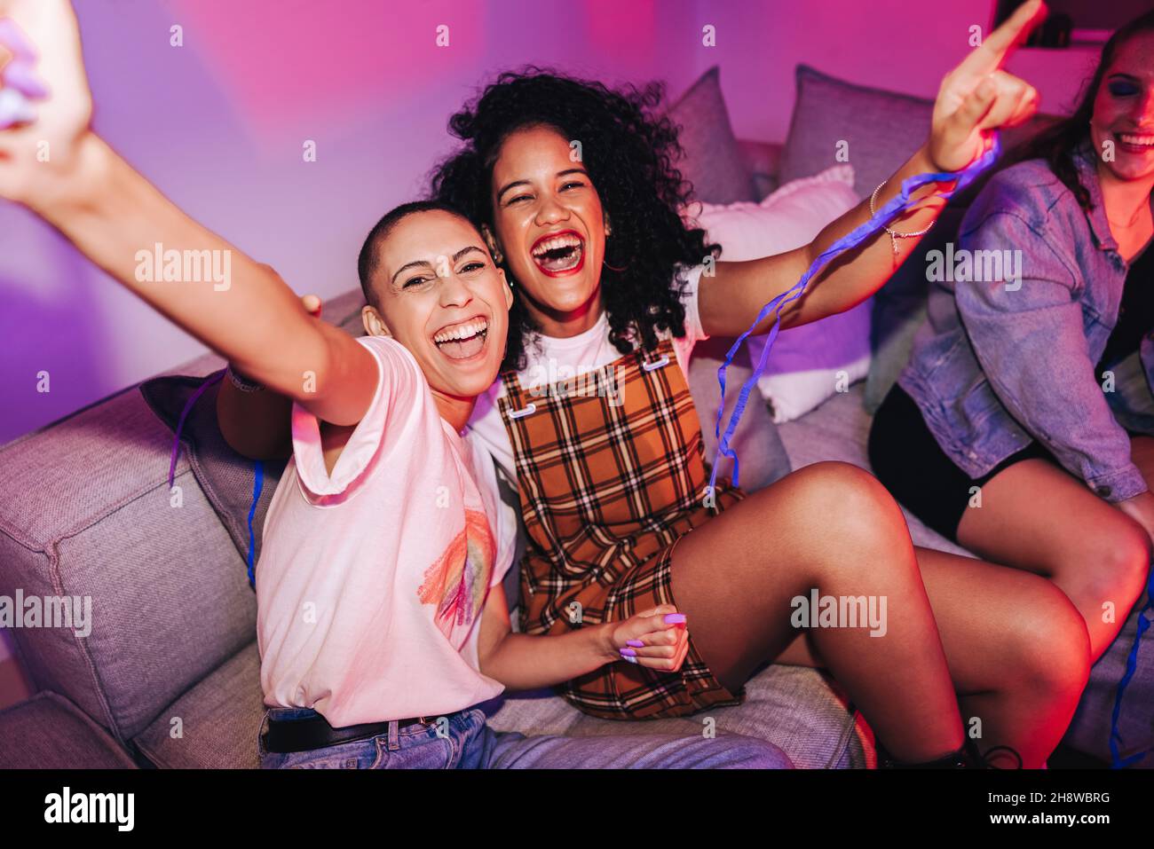 Best friends having a good time at a house party. Vibrant female friends cheering happily while sitting on a couch in neon light. Cheerful young women Stock Photo