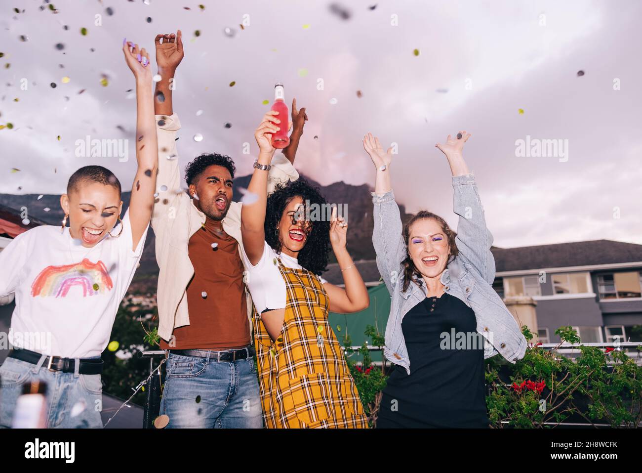 Four happy friends cheering under falling confetti. Group of generation z friends partying and celebrating on a rooftop. Multicultural friends having Stock Photo