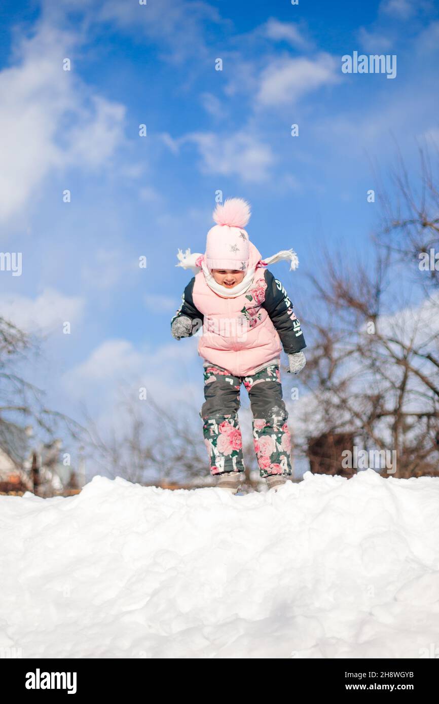 Winter landscape with blue sky. Child in warm winter suit is having fun, playing and fishing in snowdrift, riding snow slide during walk in nature on Stock Photo