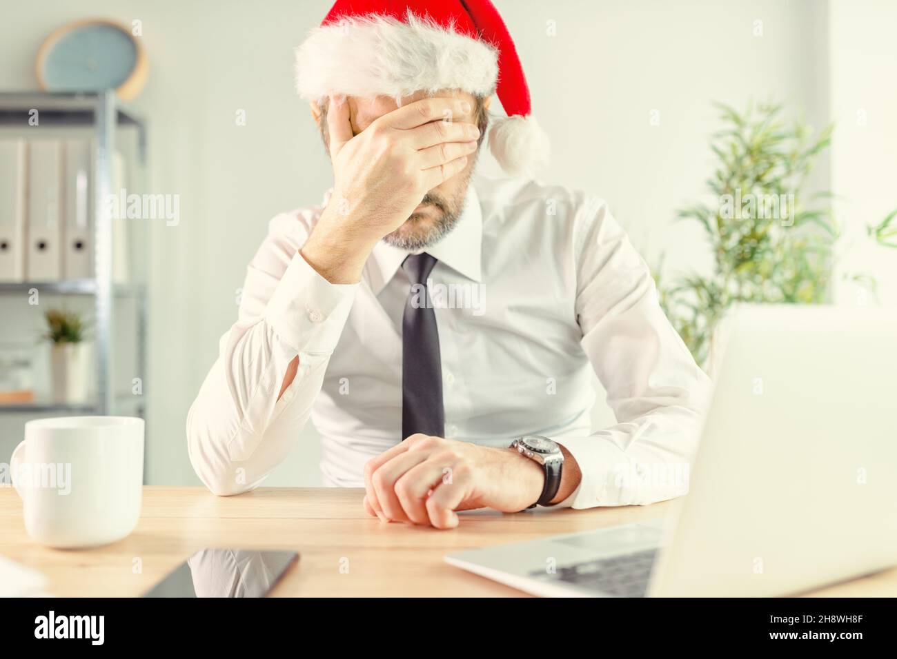 New Year depression, businessman in office wearing Santa Claus hat feeling lonely and depressed with hand covering face, selective focus Stock Photo