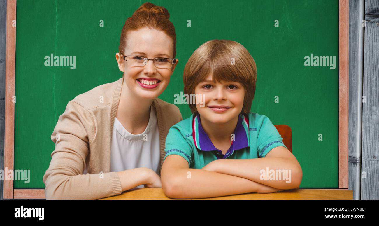 Portrait of smiling redhead teacher sitting with schoolboy against blackboard in classroom Stock Photo