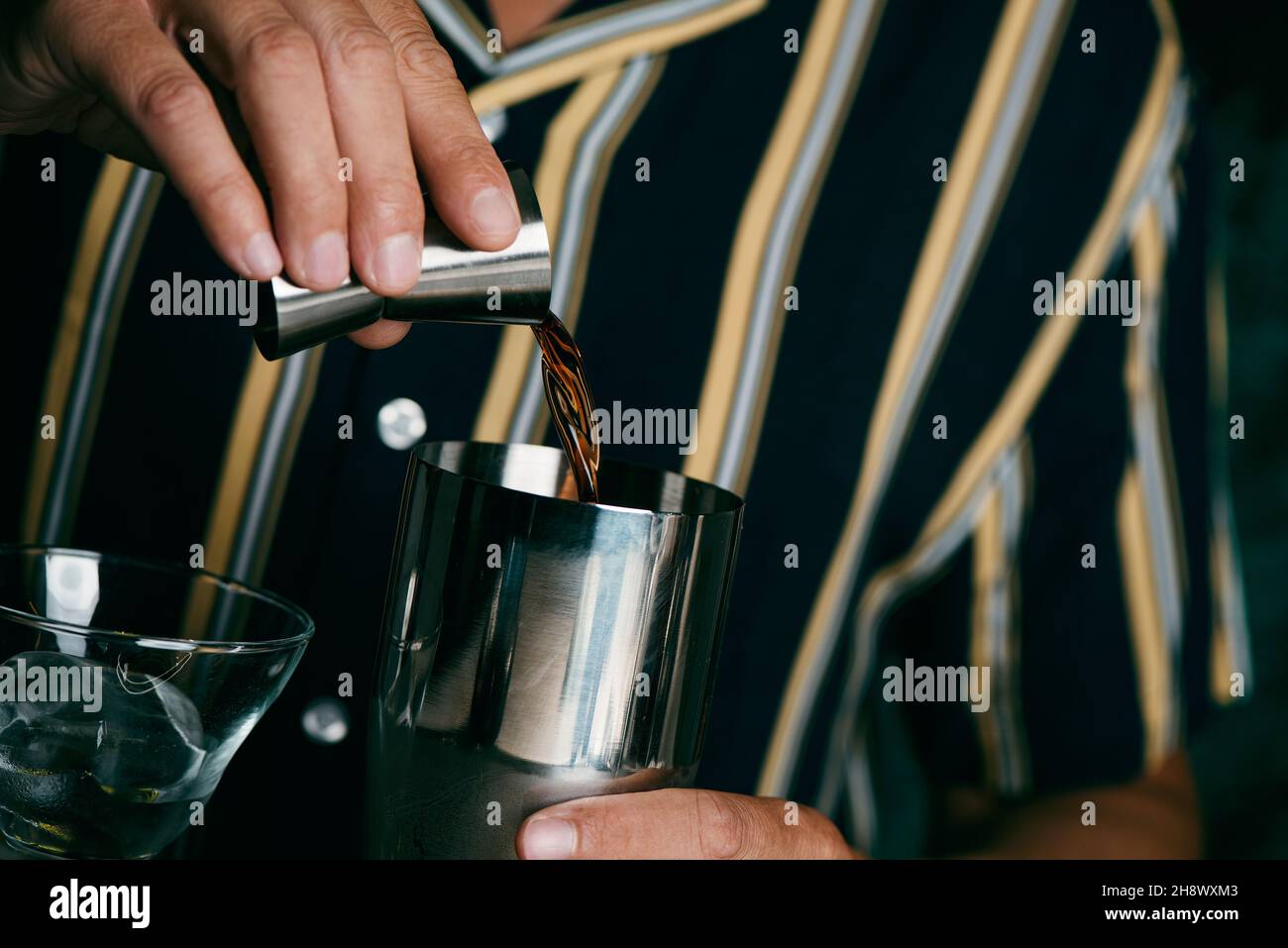 a young caucasian man, wearing a striped shirt, is about to prepare a cocktail in a metal shaker by adding a dark liquor to it with a jigger Stock Photo
