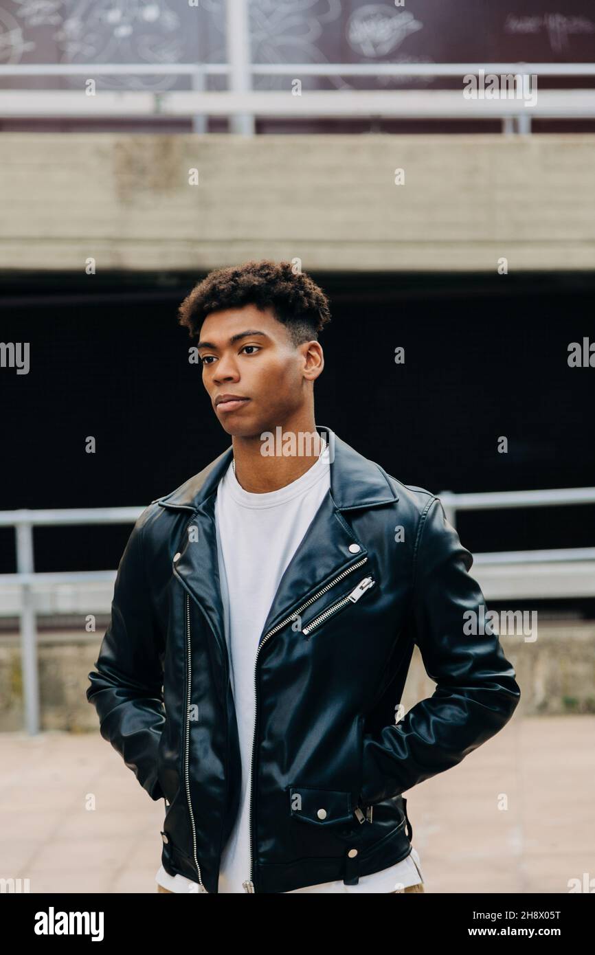 Young thoughtful black male in leather jacket standing on street with hands in pockets against railing and looking away Stock Photo