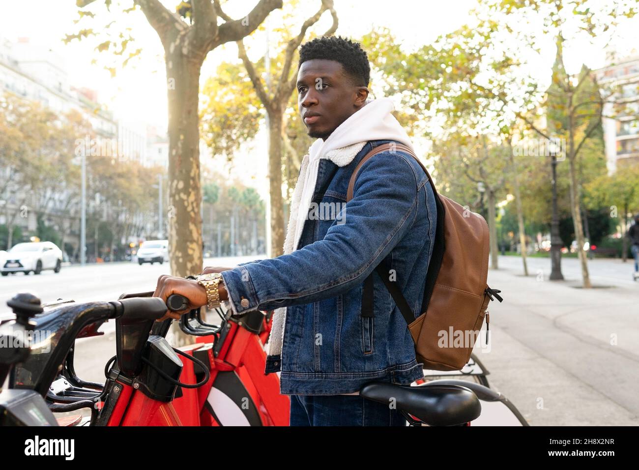 Side view of African American male with dark curly hair in denim jacket standing and taking bicycle on rental station on street in city in daylight Stock Photo