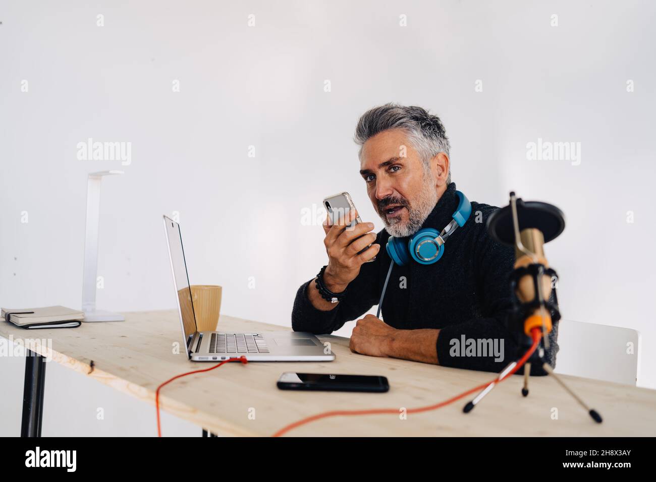 Old stylish male broadcaster with beard sitting at table with laptop and looking away while recording audio message on mobile phone Stock Photo