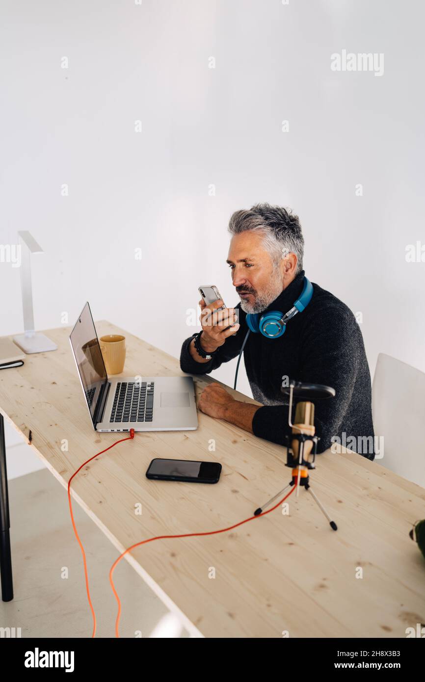 Old stylish male broadcaster with beard sitting at table with laptop and looking away while recording audio message on mobile phone Stock Photo
