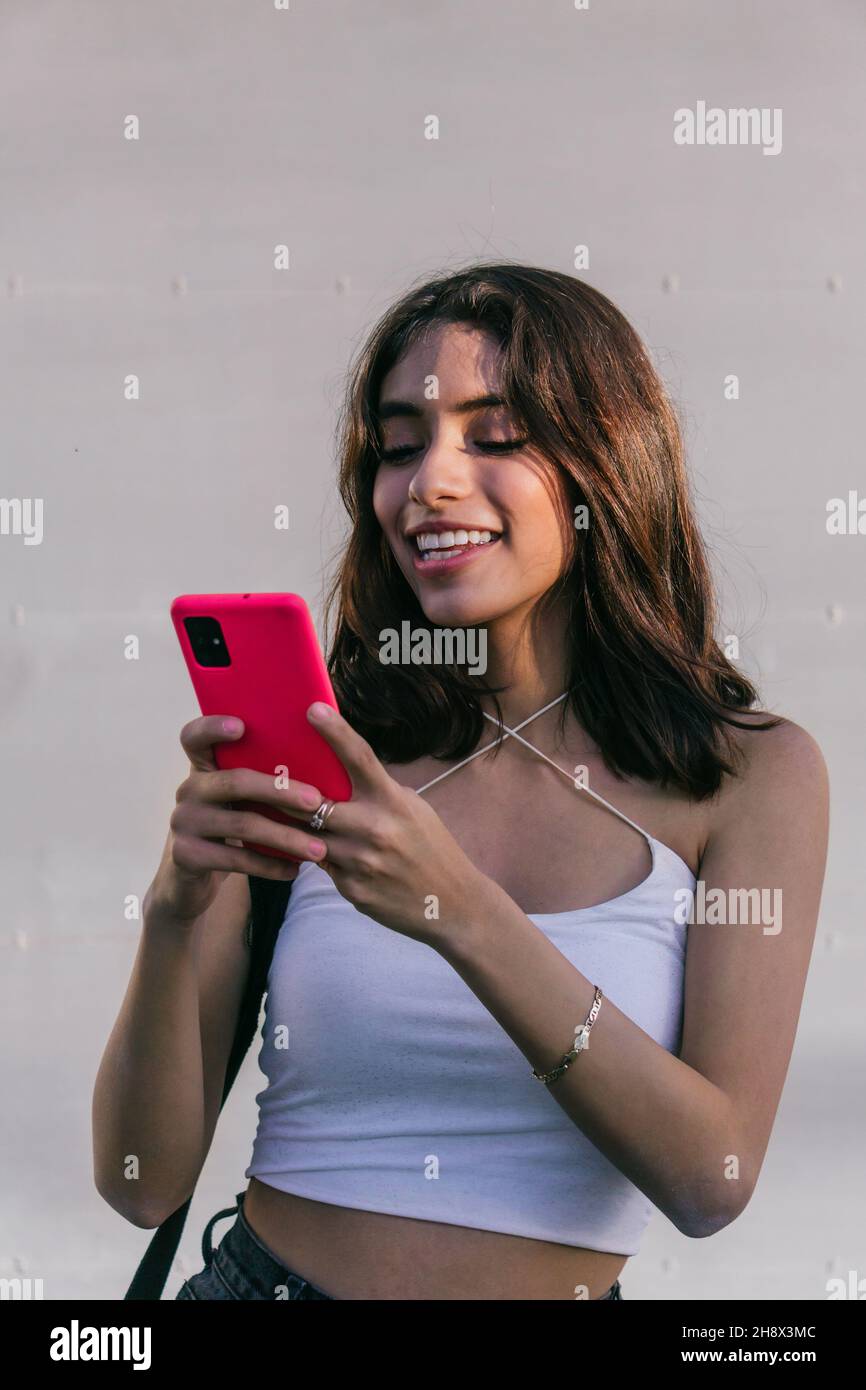 Happy Hispanic female smiling and using red cellphone while texting against white backdrop Stock Photo