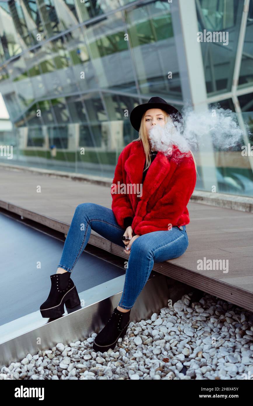 Blonde young Woman with hat and red jacket smoking with vaper machine on the street Stock Photo