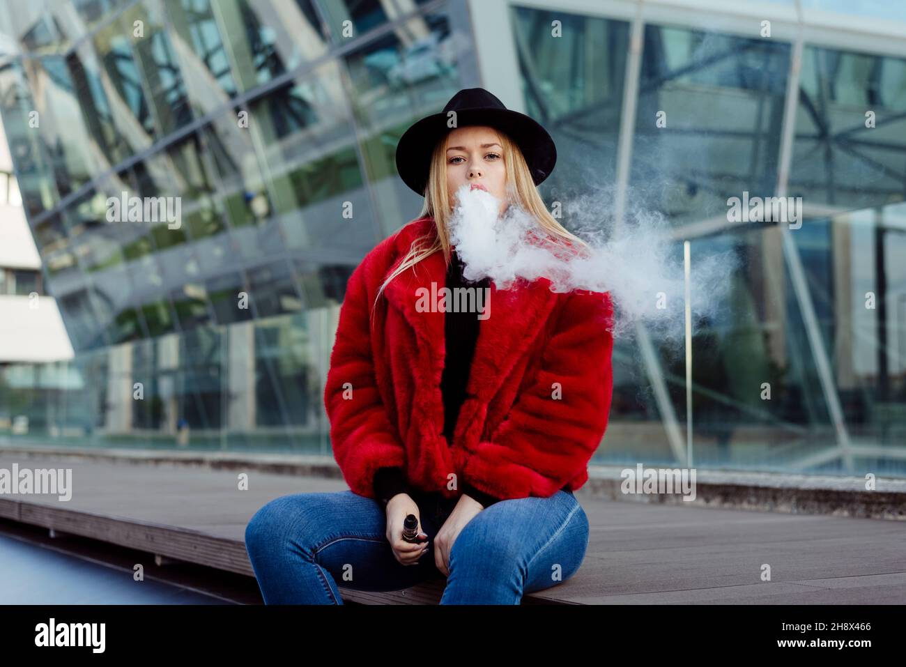 Blonde young Woman with hat and red jacket smoking with vaper machine on the street Stock Photo