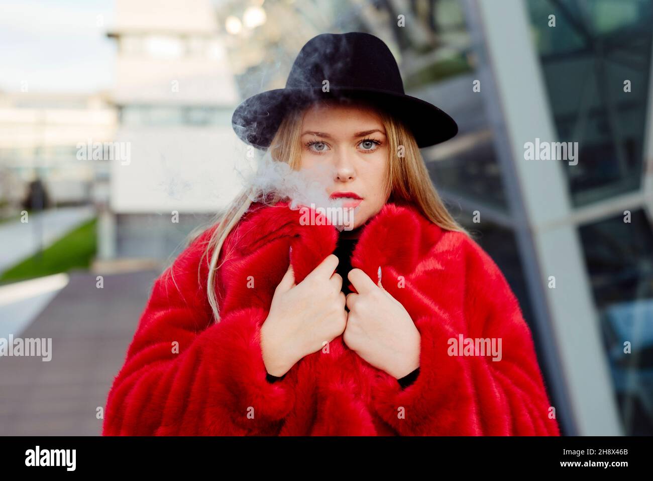 Cute blonde young Woman with hat and red jacket smoking with vaper machine on the street Stock Photo