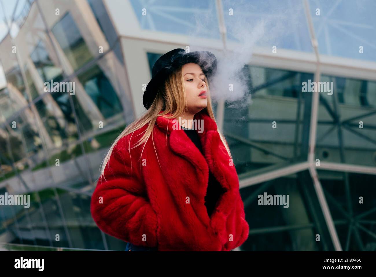 Cute blonde young Woman with hat and red jacket smoking with vaper machine on the street Stock Photo