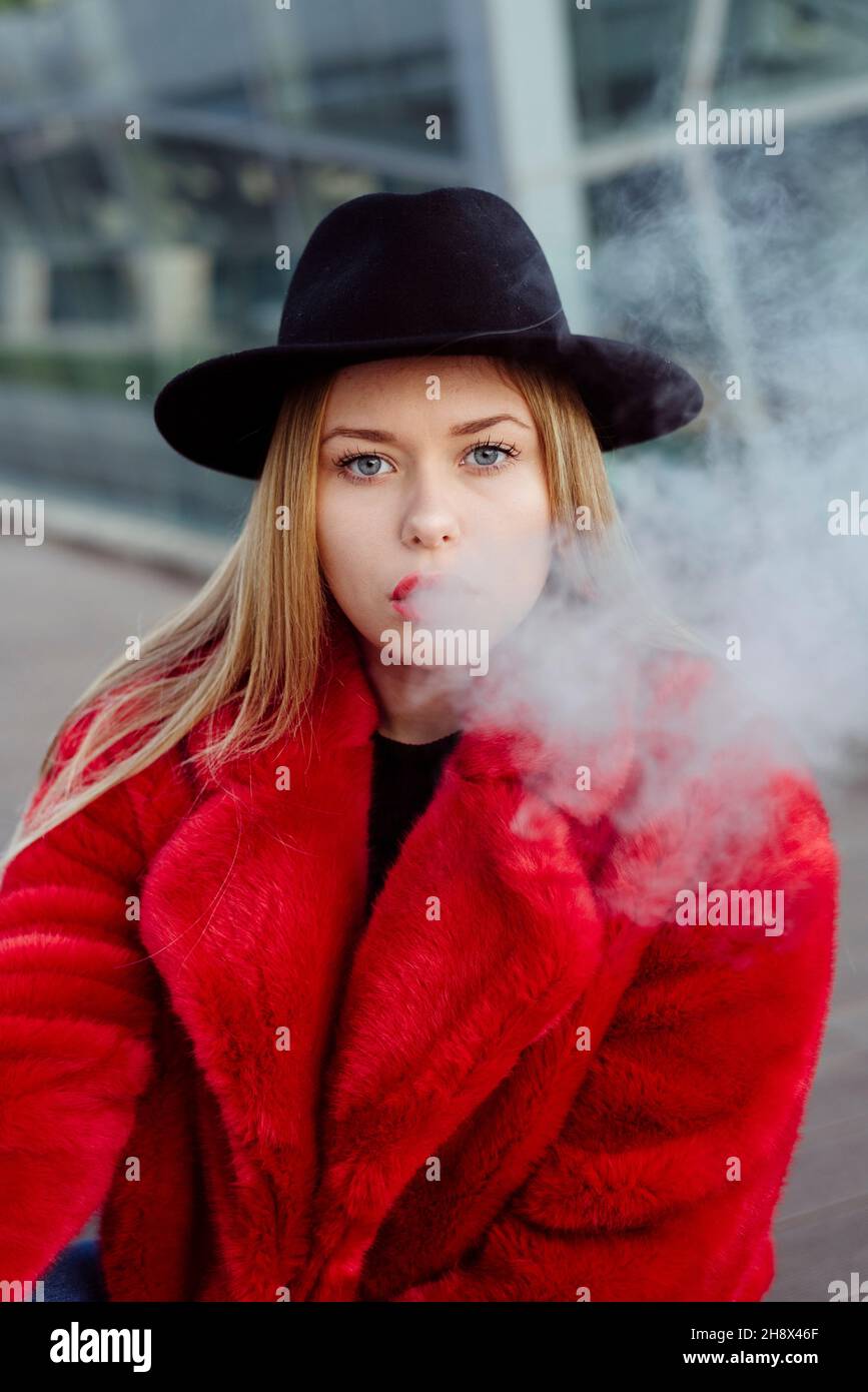 Cute blonde young Woman with hat and red jacket smoking with vaper machine on the street Stock Photo