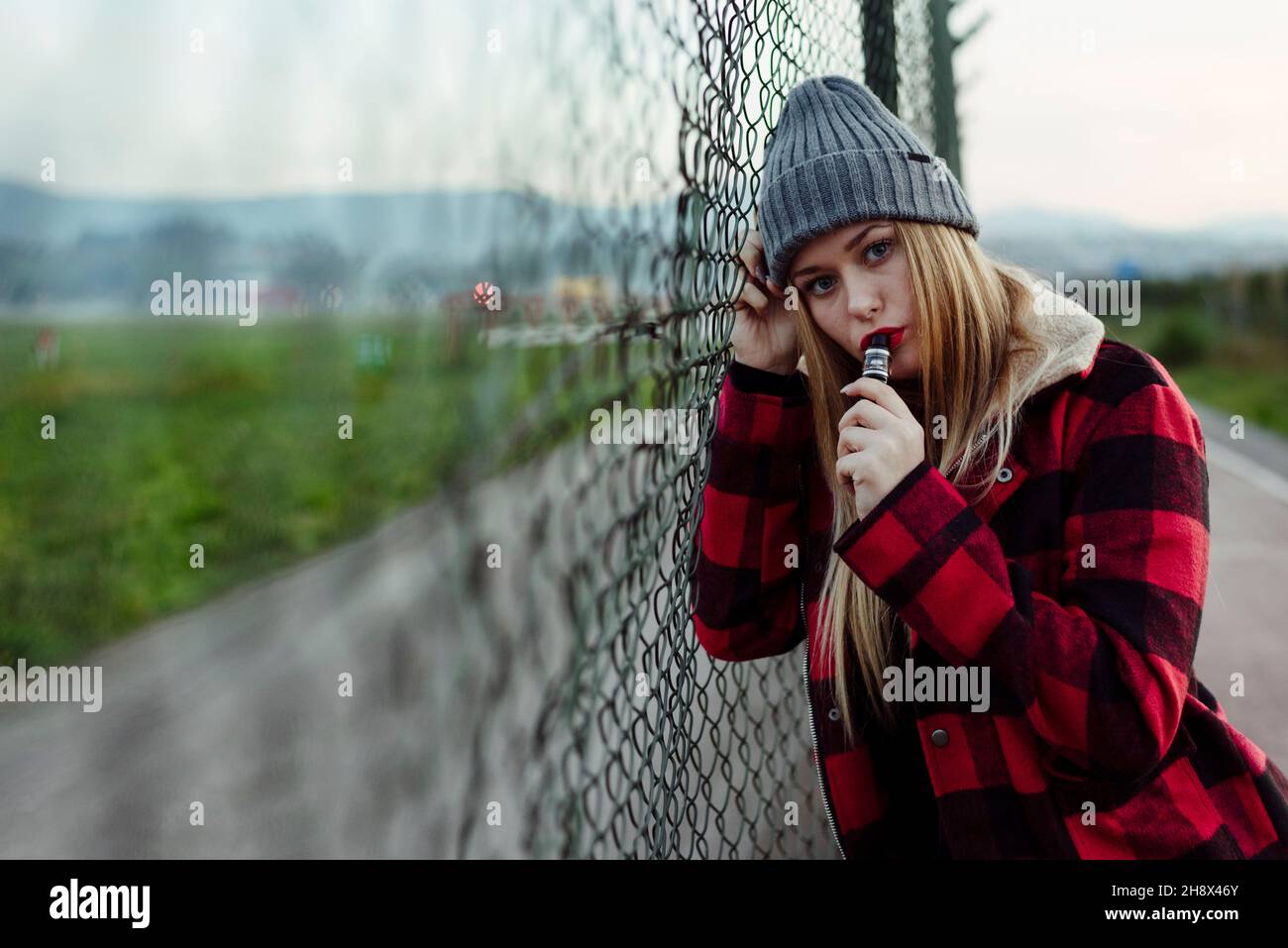 Pretty blonde young Woman with wool hat leaning on the fence and smoking with vaper machine Stock Photo